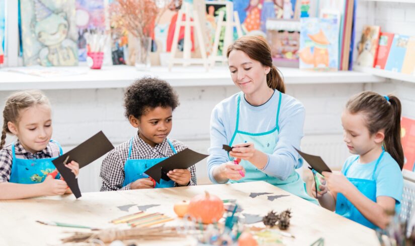 Group of intercultural schoolkids and their teacher in aprons cutting out halloween bats from sheets of black paper at lesson